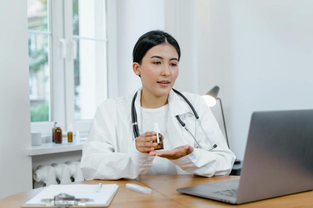 A young female doctor in a white coat on a video call, discussing medication with a patient.